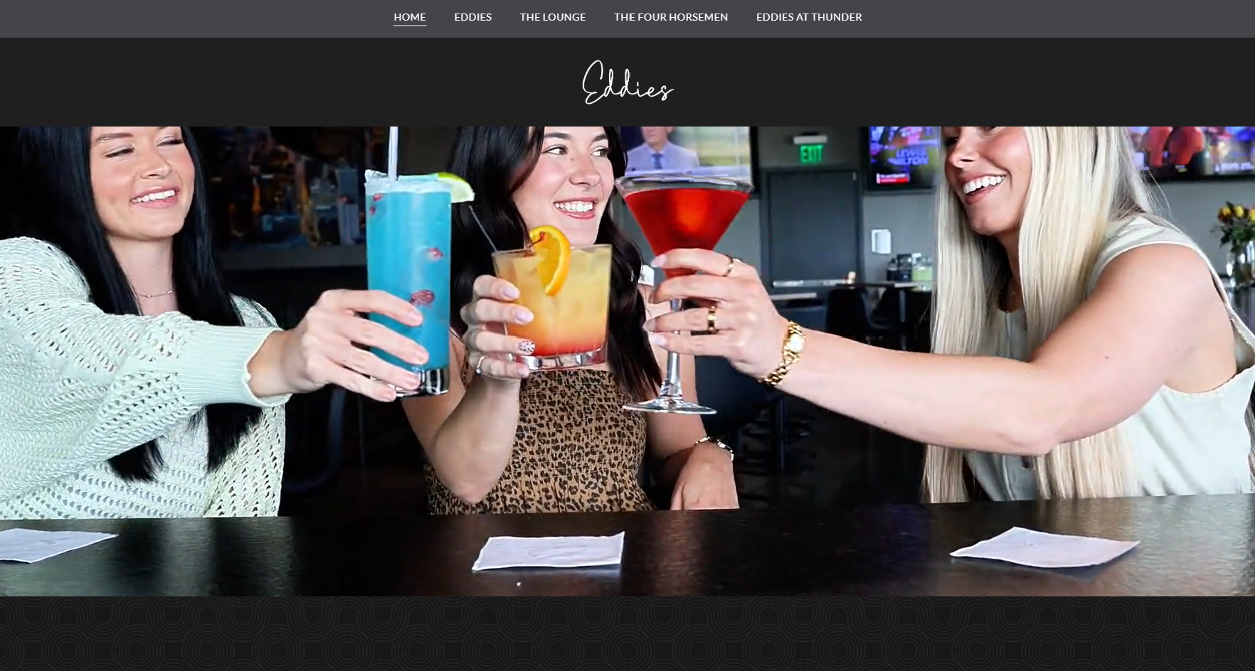 Three women at a bar toasting with colorful cocktails (blue, orange, and red) and smiling.
